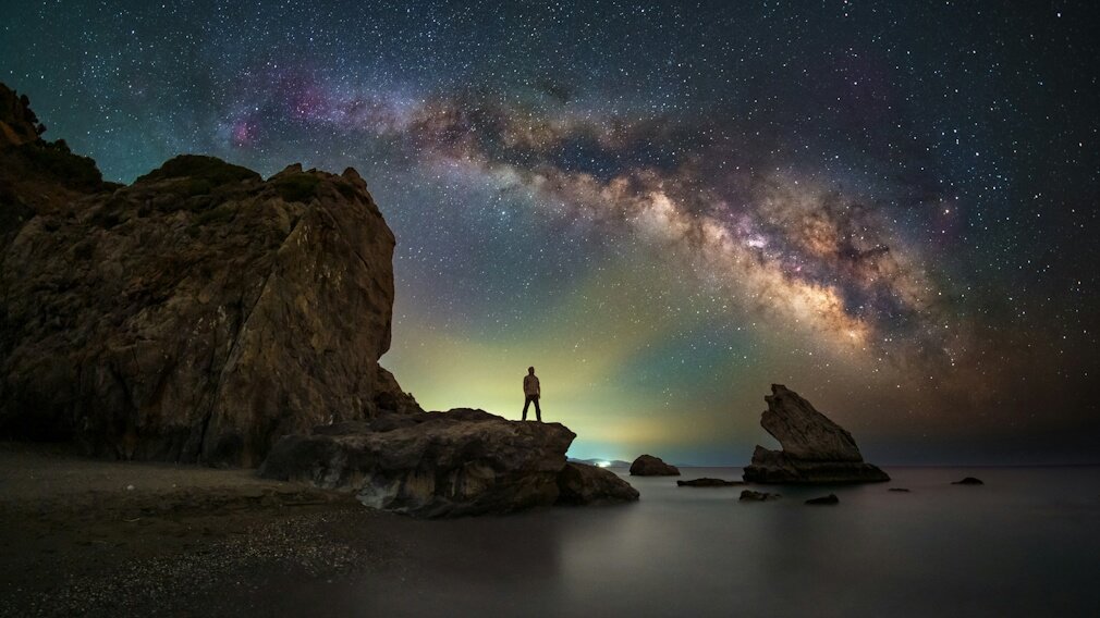 Person standing on a rocky shore looking up at the starry night sky and galaxy